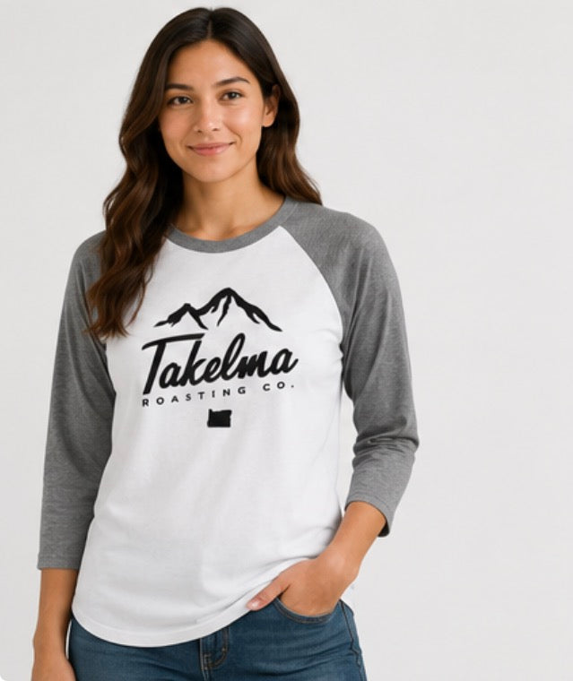 A woman wearing a Takelma Roasting Co. raglan baseball t-shirt with a white body, gray 3/4 sleeves, and a black mountain logo graphic on the chest, paired with blue jeans against a light gray background.