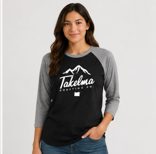 A woman modeling a black and gray raglan baseball t-shirt with a white graphic of mountains and the text 'Takelma Roasting Co.' on the front, set against a plain light gray background.