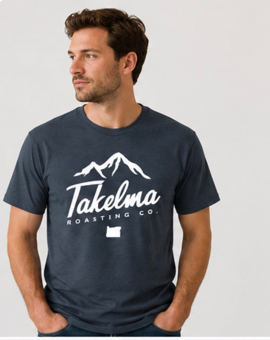 A man wearing a navy blue crewneck t-shirt with a white graphic print that reads 'Takelma Roasting Co.' with a mountain illustration and an Oregon state silhouette, set against a plain light gray background.