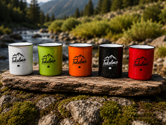 Five enamel camping mugs in white, lime green, orange, black, and red, each printed with the black Takelma Mountain Co. logo, are lined up on a rustic log outdoors with a blurred mountain stream and forest background.